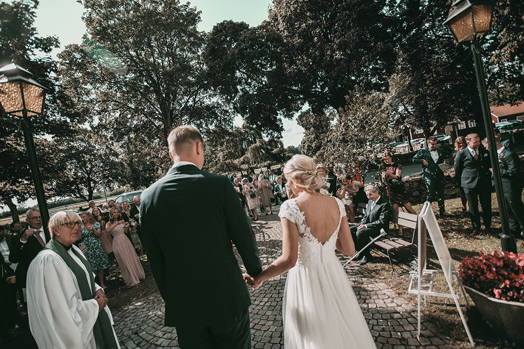 Brudparet möter glada gäster utanför kyrkan i Tånnö efter vigseln. En strålande brud i vit klänning och blomsterbukett tar sin nygifta make i handen, omgivna av jublande vänner och familj