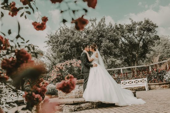 Ett brudpar som kysser varandra i de pittoreska Sofieros trädgårdar. I förgrunden sträcker sig livfulla röda blommor ut, som framhäver parets kärleksfulla kyss. Det är en dynamisk och passionerad bild som fångar ett intimt ögonblick mot en bakgrund av naturlig skönhet och färg.