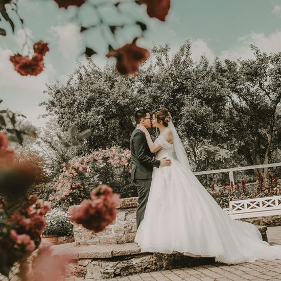 Ett brudpar som kysser varandra i de pittoreska Sofieros trädgårdar. I förgrunden sträcker sig livfulla röda blommor ut, som framhäver parets kärleksfulla kyss. Det är en dynamisk och passionerad bild som fångar ett intimt ögonblick mot en bakgrund av naturlig skönhet och färg.